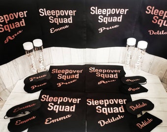 May include: Black sleep masks with the text "Sleepover Squad" and names "Emma", "Delilah", "Prue", and "Ana" in rose gold lettering.  Black sleep shirts with the same text and names.  Clear water bottles with names in white lettering.