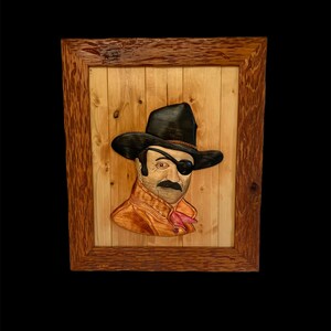 May include: A wooden framed artwork featuring a carved wooden portrait of a man wearing a black hat and an eyepatch. The man has a mustache and beard. The background is a light wood.