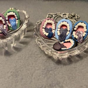 May include: A collection of handmade clay pendants shaped like open mouths, each with a unique color scheme. The pendants are displayed in clear, heart-shaped glass dishes. The mouths feature white teeth, pink gums, and colorful tongues.