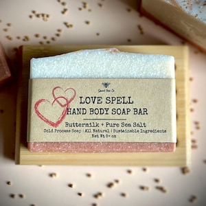 May include: A rectangular Love Spell hand and body soap bar with a white top layer and a pink bottom layer, wrapped in a tan paper label featuring two red hearts, the text Love Spell Hand Body Soap Bar, and a net weight of 5+ oz.
