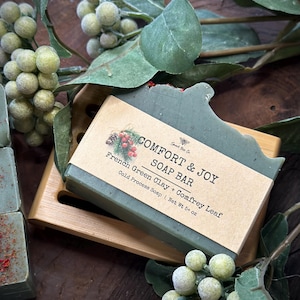 May include: A green soap bar with a brown label that reads "COMFORT & JOY SOAP BAR French Green Clay + Comfrey Leaf Cold Process Soap | Net Wt 5+ oz".