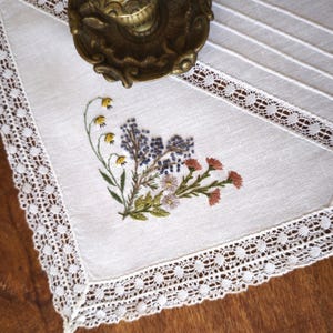Peut inclure: Un chemin de table blanc orné d'une bordure en dentelle blanche complexe et d'un bouquet floral brodé au centre. La broderie présente des fleurs jaunes, bleues et roses avec des tiges et des feuilles vertes. Un objet métallique est posé au-dessus.