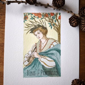 Może przedstawiać: Akwarelowa ilustracja na papierze zatytułowana King of Pentacles, przedstawiająca postać w wieńcu laurowym grającą na flecie pod drzewem pomarańczowym. Postać ma na sobie kremową tunikę ze złotymi wykończeniami oraz turkusowy płaszcz.