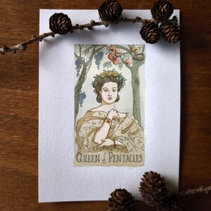 May include: A rectangular art print featuring a watercolor illustration of the Queen of Pentacles tarot card. The figure wears a yellow floral crown and a patterned shawl, surrounded by grapevines, fruit trees, and a small bird in a nest.