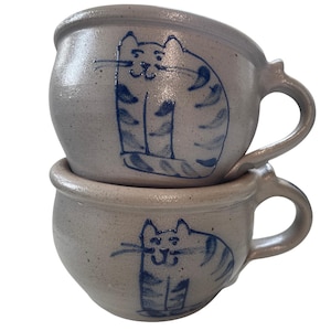 May include: Two stacked ceramic mugs with a speckled light gray finish. Each mug features a hand-painted dark blue line drawing of a stylized cat with stripes, whiskers, and a simple facial expression. The mugs have rounded bodies and side handles.