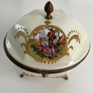 May include: A cream-coloured porcelain trinket box with a hinged lid and a brass finial. The lid features a central hand-painted scene of a person playing a bagpipe for another person, surrounded by gold filigree and a decorative brass metal rim.