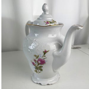 May include: A white porcelain teapot featuring a delicate floral pattern of pink roses and green leaves. The teapot has a scalloped rim with a gold-tone trim, an embossed scroll design on the body, a curved handle, and a matching lid with a finial.