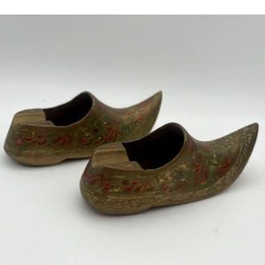 May include: A pair of brass shoe-shaped decorative objects featuring an etched floral pattern with red and green enamel accents. Each piece has a pointed toe and a hollow interior, showcasing a metallic finish with intricate engraved detailing.