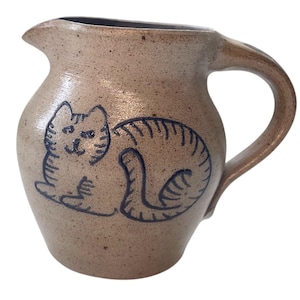 May include: A tan speckled ceramic pitcher featuring a dark blue hand-drawn illustration of a sitting tabby cat. The pitcher has a rounded body, a curved handle on the right side, and a small pouring spout at the top rim of the vessel.