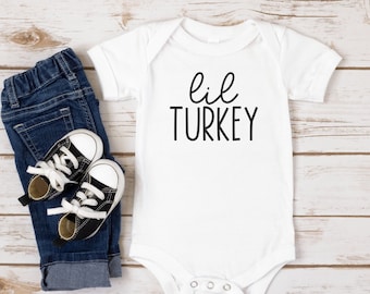 May include: A white baby onesie with the text "lil TURKEY" in black script. The onesie is paired with a pair of dark blue denim jeans and small black and white sneakers. The items are arranged on a light wood-grain surface.