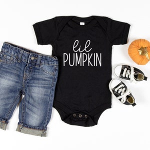 Puede incluir: Un body negro para bebé con el texto blanco "lil PUMPKIN", junto con pantalones cortos de mezclilla azules y zapatillas deportivas pequeñas en blanco y negro. También hay una decoración de calabaza naranja.