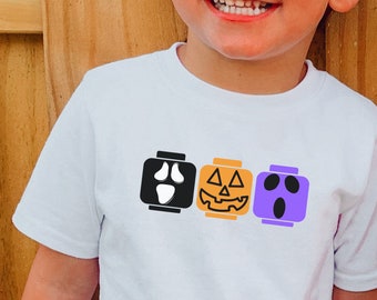 May include: A white t-shirt with a design of three LEGO bricks. The bricks are decorated with Halloween-themed faces: a ghost, a pumpkin, and another ghost.
