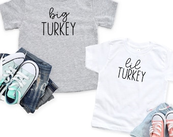 May include: Two t-shirts for kids, one gray and one white, with the text "big TURKEY" and "lil TURKEY" printed on them. The shirts are laid out on a white background with a pair of blue jeans and two pairs of sneakers.