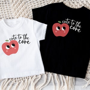 K&ouml;nnte beinhalten: Zwei T-Shirts mit einem roten Apfel-Motiv und dem Text "cute to the core". Ein T-Shirt ist wei&szlig; und das andere ist schwarz.