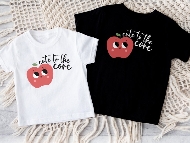 K&ouml;nnte beinhalten: Zwei T-Shirts mit einem roten Apfel-Motiv und dem Text "cute to the core". Ein T-Shirt ist wei&szlig; und das andere ist schwarz.