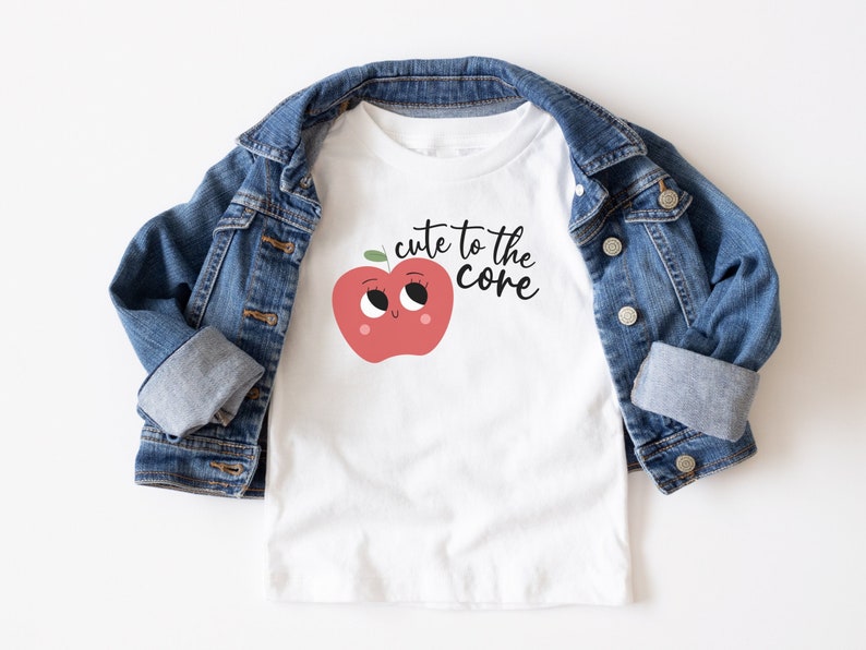 K&ouml;nnte beinhalten: Wei&szlig;es T-Shirt mit einem roten Apfel-Motiv und dem Text "cute to the core" in Schwarz. Das Shirt ist gefaltet und liegt auf einer blauen Jeansjacke.