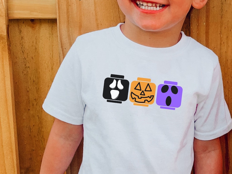 Op de afbeelding: Een wit T-shirt met een ontwerp van drie LEGO-stenen. De stenen zijn versierd met Halloween-gezichten: een spook, een pompoen en nog een spook.