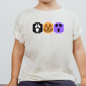 Op de afbeelding: Een wit T-shirt met een zwarte geest, een oranje pompoen en een paars spookgezicht erop gedrukt. De gezichten zijn in de vorm van Lego-blokken.