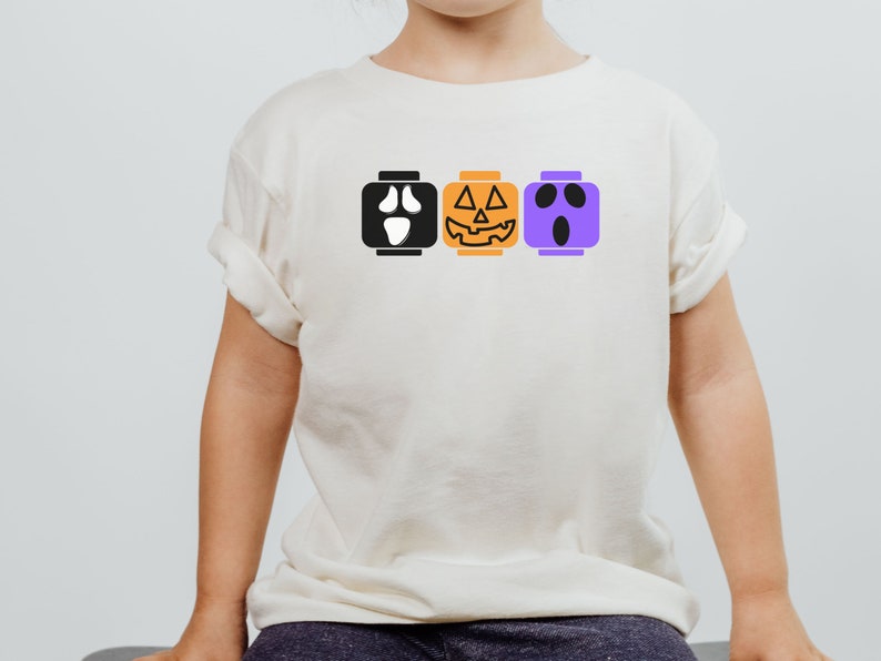 Op de afbeelding: Een wit T-shirt met een zwarte geest, een oranje pompoen en een paars spookgezicht erop gedrukt. De gezichten zijn in de vorm van Lego-blokken.