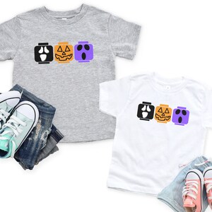 Op de afbeelding: Twee kinder T-shirts met een Halloween-thema. De shirts hebben een rij van drie Lego-achtige blokken met verschillende Halloween-gezichten: een spook, een pompoen en nog een spook. De shirts zijn grijs en wit.