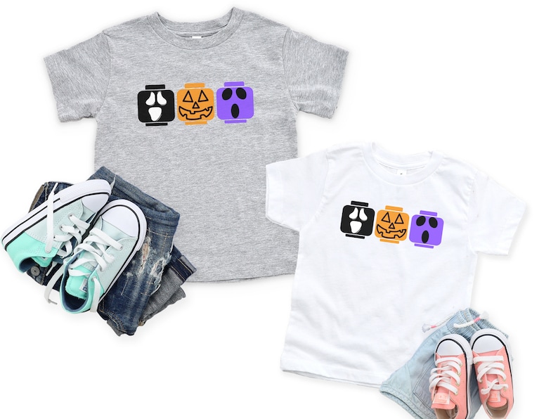 Op de afbeelding: Twee kinder T-shirts met een Halloween-thema. De shirts hebben een rij van drie Lego-achtige blokken met verschillende Halloween-gezichten: een spook, een pompoen en nog een spook. De shirts zijn grijs en wit.