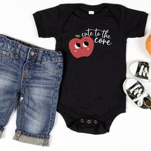 K&ouml;nnte beinhalten: Ein schwarzer Baby-Strampler mit einem roten Apfel-Motiv und dem Text "cute to the core". Der Strampler wird mit blauen Jeans-Shorts und schwarzen High-Top-Sneakern kombiniert.