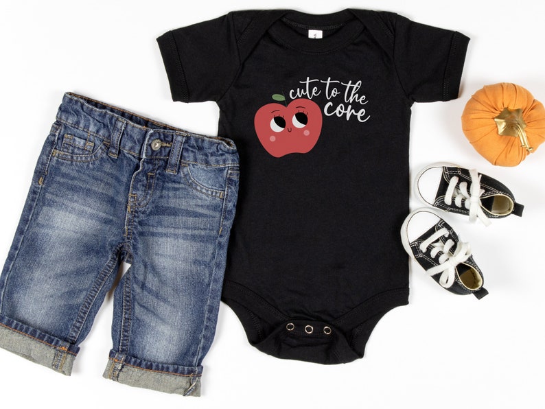 K&ouml;nnte beinhalten: Ein schwarzer Baby-Strampler mit einem roten Apfel-Motiv und dem Text "cute to the core". Der Strampler wird mit blauen Jeans-Shorts und schwarzen High-Top-Sneakern kombiniert.