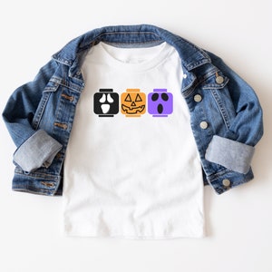 Op de afbeelding: Een wit T-shirt met een ontwerp van drie Halloween-Lego-blokken. De blokken tonen een spook, een pompoen en een monster.
