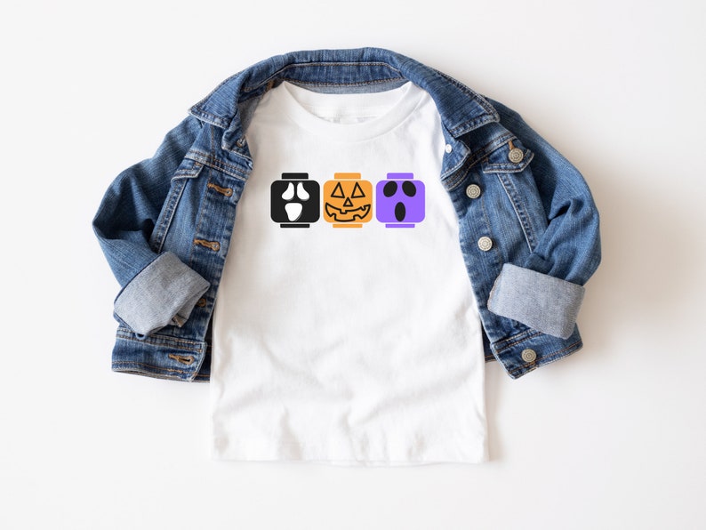 Op de afbeelding: Een wit T-shirt met een ontwerp van drie Halloween-Lego-blokken. De blokken tonen een spook, een pompoen en een monster.