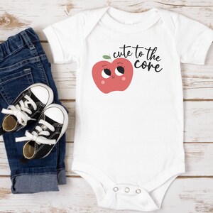 K&ouml;nnte beinhalten: Wei&szlig;er Baby-Strampler mit einem roten Apfel-Motiv und dem Text "cute to the core". Der Strampler wird mit einer blauen Jeans und schwarzen Sneakern kombiniert.
