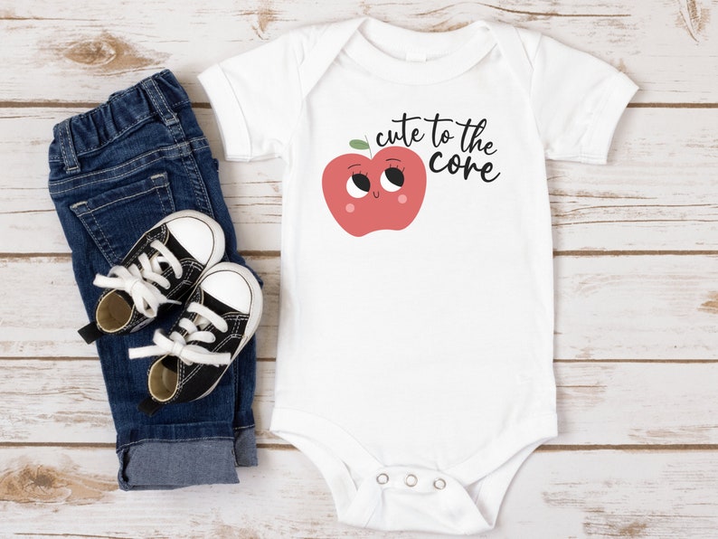 K&ouml;nnte beinhalten: Wei&szlig;er Baby-Strampler mit einem roten Apfel-Motiv und dem Text "cute to the core". Der Strampler wird mit einer blauen Jeans und schwarzen Sneakern kombiniert.