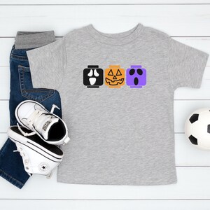 Op de afbeelding: Een grijs T-shirt met een ontwerp van drie Halloween-Lego-blokken. De blokken zijn een spook, een pompoen en een Frankenstein. Het shirt wordt gecombineerd met een blauwe jeans en witte sneakers.