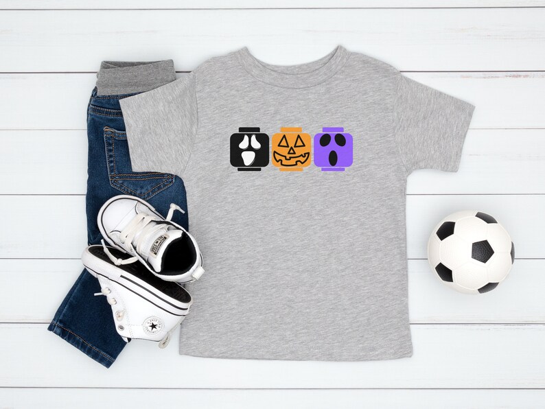 Op de afbeelding: Een grijs T-shirt met een ontwerp van drie Halloween-Lego-blokken. De blokken zijn een spook, een pompoen en een Frankenstein. Het shirt wordt gecombineerd met een blauwe jeans en witte sneakers.