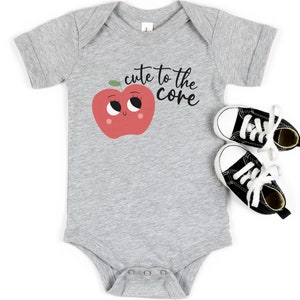 K&ouml;nnte beinhalten: Ein grauer Baby-Strampler mit einem roten Apfel-Motiv und dem Text "cute to the core".