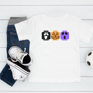 Op de afbeelding: Een wit T-shirt met een ontwerp van drie Halloween-Lego-blokken. De blokken zijn een spook, een pompoen en een monster. Het shirt ligt op een wit houten oppervlak met een paar witte sneakers en een blauwe jeans.