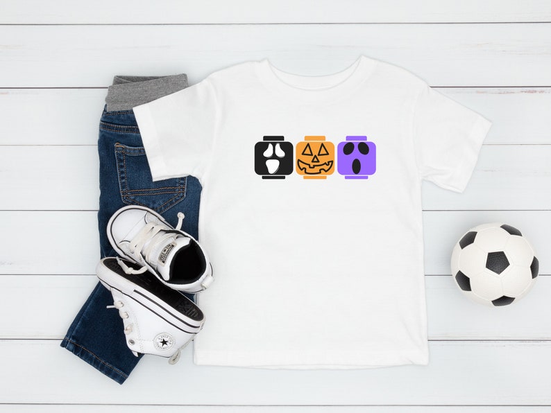 Op de afbeelding: Een wit T-shirt met een ontwerp van drie Halloween-Lego-blokken. De blokken zijn een spook, een pompoen en een monster. Het shirt ligt op een wit houten oppervlak met een paar witte sneakers en een blauwe jeans.