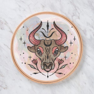 Peut inclure: Un cercle de broderie au point de croix représentant une tête de taureau stylisée avec des cornes roses et des accents floraux. Le motif est placé sur un fond de style aquarelle aux couleurs pastel. Le cercle est en bois.