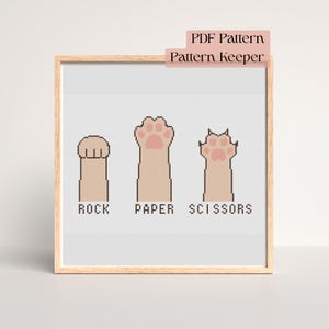 Puede incluir: Un bordado a punto de cruz enmarcado que representa un juego de Piedra, Papel, Tijeras con patas de gato. Las patas son beige con almohadillas rosas, y las palabras "ROCK", "PAPER" y "SCISSORS" están debajo. El marco es marrón claro.