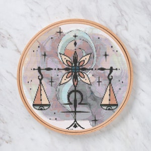 May include: A round, wooden embroidery hoop holds a cross-stitch design of the Libra zodiac symbol. The design features a scale, a floral element, and a watercolor-style background in pastel colors, including peach, blue, and gray.