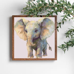May include: A framed cross-stitch of a baby elephant. The elephant is depicted in shades of blue, tan, and gray, with large ears and a curled trunk. The artwork is set against a light pink background and framed in a brown wooden frame.
