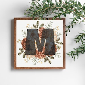 Op de afbeelding: Een ingelijste kruissteek met de letter "M" in een donker bloemenpatroon, omringd door een krans van perzik- en crèmekleurige bloemen en groene bladeren. Het frame is van hout.