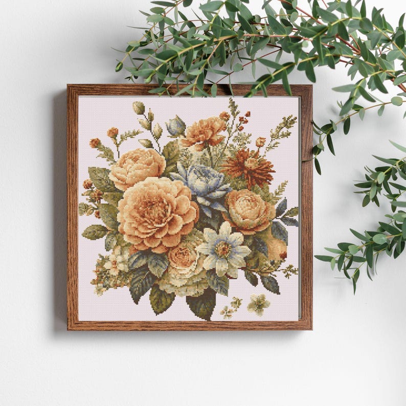 K&ouml;nnte beinhalten: Ein gerahmtes Kreuzstich-Blumenbouquet mit gro&szlig;en, goldgelben Rosen, blauen und wei&szlig;en Blumen und verschiedenen Gr&uuml;npflanzen. Das Kunstwerk ist vor einem hellrosa Hintergrund platziert und in dunklem Holz gerahmt.