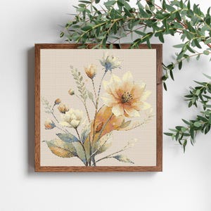 以下が含まれることがあります： 額装された花のクロスステッチのデザイン。このアートワークは、黄色と白の花のブーケと緑と青の葉が特徴です。フレームは茶色の木でできており、背景は明るいベージュ色です。