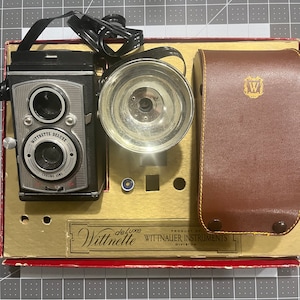 May include: A vintage black and silver Wittnitte Deluxe camera with a flash attachment and a brown leather case. The camera has a chrome lens and a black strap. The case has a gold W logo.