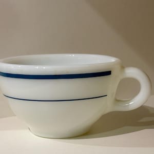 May include: White ceramic teacup with a rounded body and a handle. The cup is decorated with two horizontal blue stripes. The cup is sitting on a white surface.