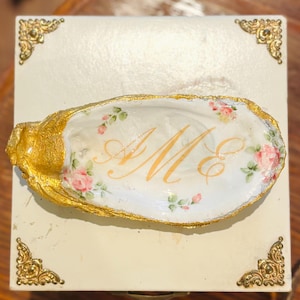 May include: A decorative oyster shell dish with a gold-painted rim and the initials "AM" in gold script. The shell is adorned with pink roses and green leaves. The dish rests on a cream-colored box with gold corner accents.