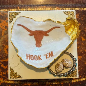 May include: A decorative oyster shell dish with a gold-painted edge, featuring a burnt orange longhorn logo and the words "HOOK 'EM". A bracelet with turquoise accents and pearl earrings are next to the dish.