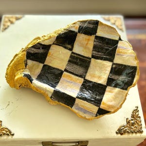 May include: A decorative oyster shell dish featuring a hand-painted black and white checkerboard pattern on the interior surface. The outer edges and the rim of the shell are finished with a textured, metallic gold paint application.