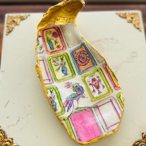May include: A decorative oyster shell dish with a gold-painted edge. The interior features a colorful, hand-painted design with framed images of flowers, a peacock, and other whimsical elements. The dish is set on a white surface.