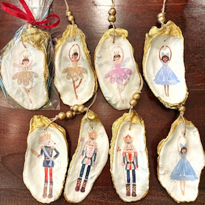 May include: Hand-painted oyster shell ornaments featuring ballet dancers and nutcracker figures. Each shell has a gold-painted edge and is strung with a gold cord and beads. The ornaments are in various colors, including pink, blue, and red.